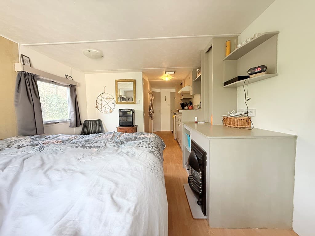 Interieur van een woning met een bed, kitchenette en een zithoek, lichte muren en houten vloer.