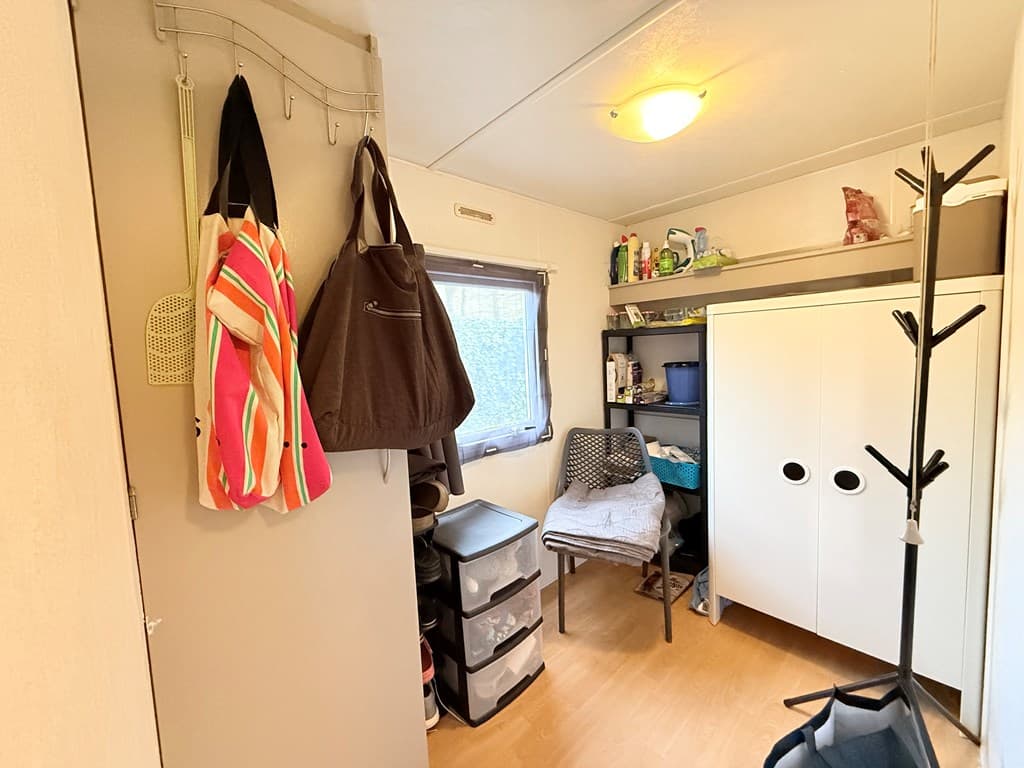 In een compacte ruimte met een kast, planken en opbergdozen, jassen en een stoel hangen aan de muur