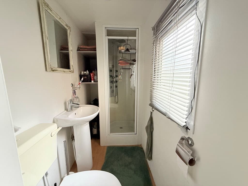 Badkamer met douchecabine, wastafel en toilet, voorzien van een raam met jaloezieën en een spiegel aan de wand