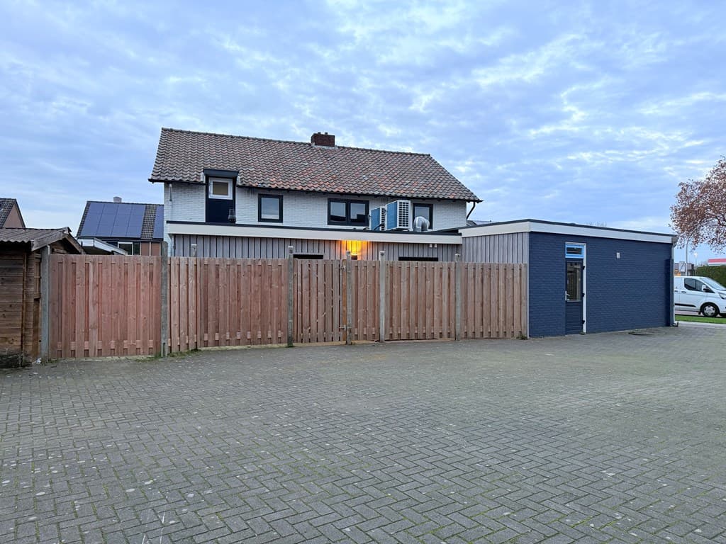 Twee verdiepingen tellend pand met schuin dak en aanbouw, omringd door een hek en een verharde parkeerplaats.