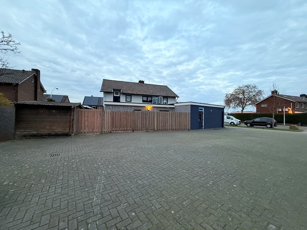 Op de foto is een parkeerplaats zichtbaar met een houten omheining en een bedrijfsgebouw op de achtergrond bij een bewolkte l