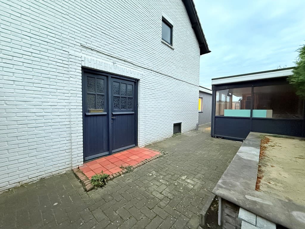 Zijaanzicht van een wit gebouw met een dubbele deur en een betegeld pad richting een overdekte ruimte.