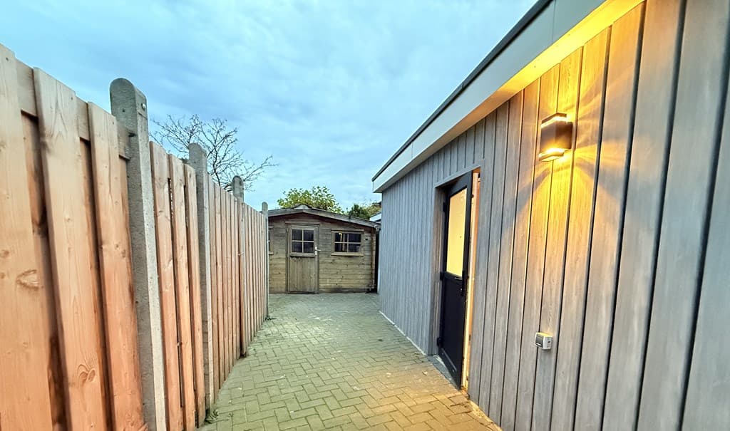 Smalle looppad met houten schutting, toegang tot woning met buitenverlichting en schuur op de achtergrond.