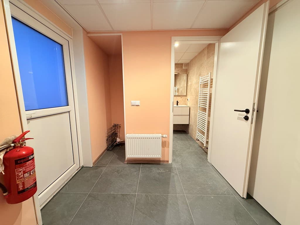 Ingangsruimte met lichtgekleurde muren, tegels op de vloer, en toegang tot een ruimte met sanitair aan de achterkant.