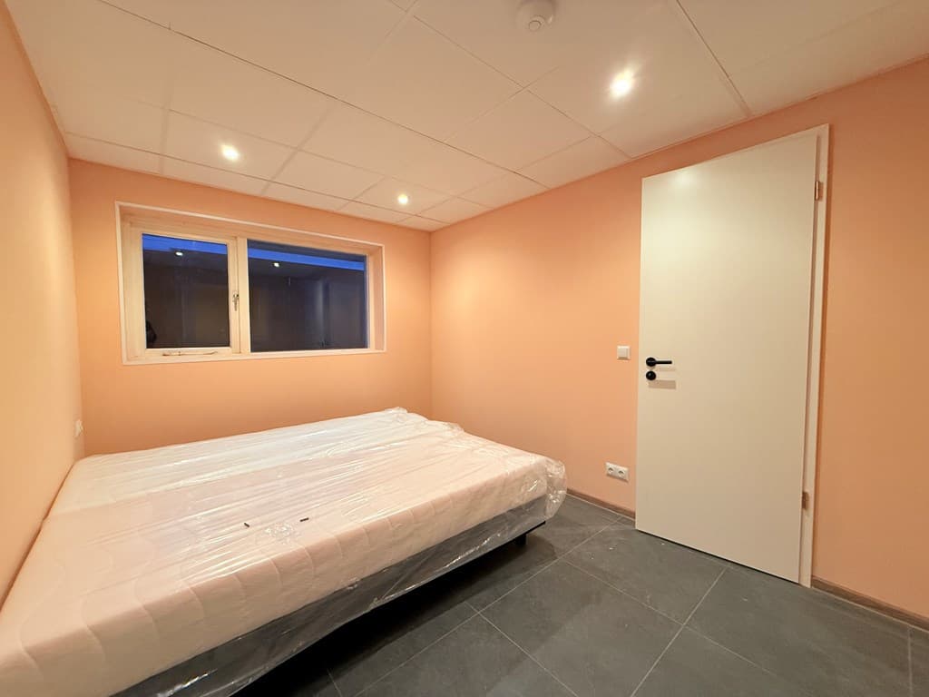 Slaapkamer met een lege matras op een keramische vloer en een raam met blauw licht aan de achterkant.