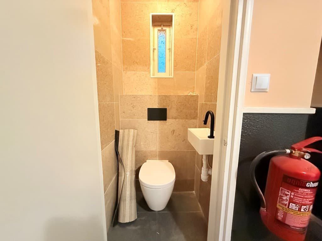 Klein toilet met tegelwanden, wastafel en een bovenraam, naast een brandblusser en een opgerolde mat.