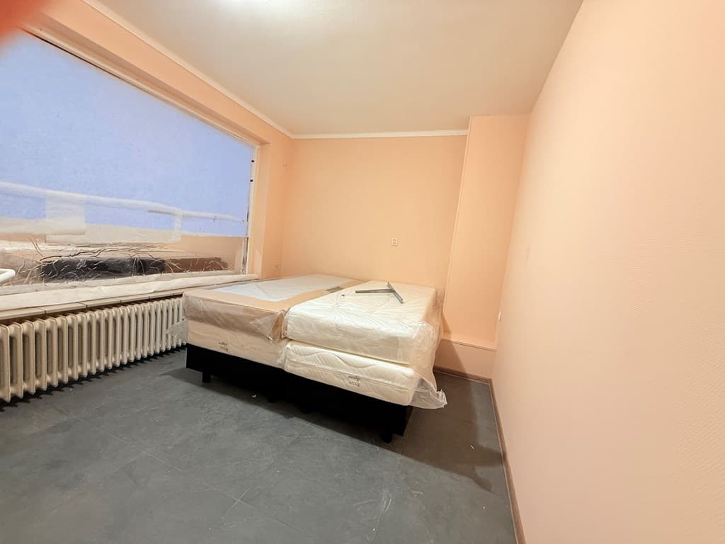 Slaapkamer met twee matrassen op een bed en een groot raam aan de ene zijde met een pastelroze wand.