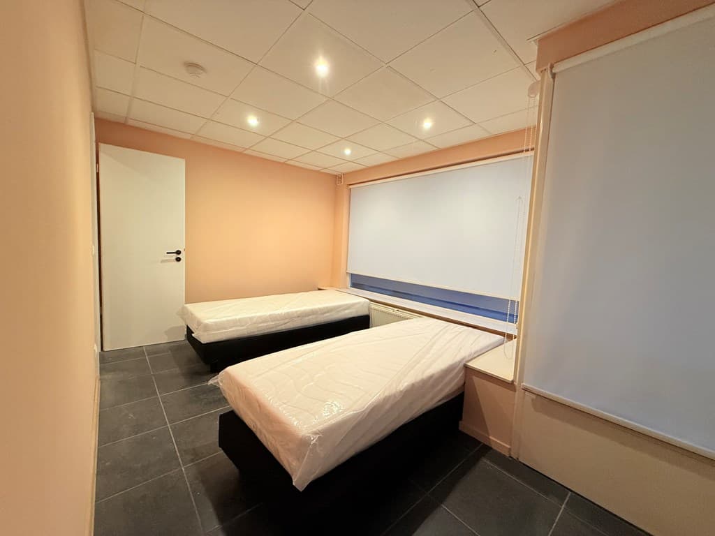 Kamer met twee eenpersoonsbedden, lichte muren en een groot raam met een rolhor.