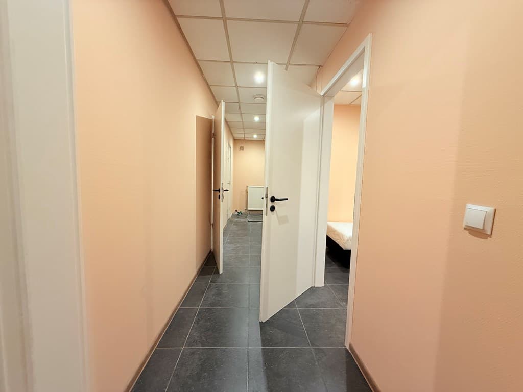 Een gang met lichtgekleurde muren en donkere tegels, met open deuren naar verschillende kamers.