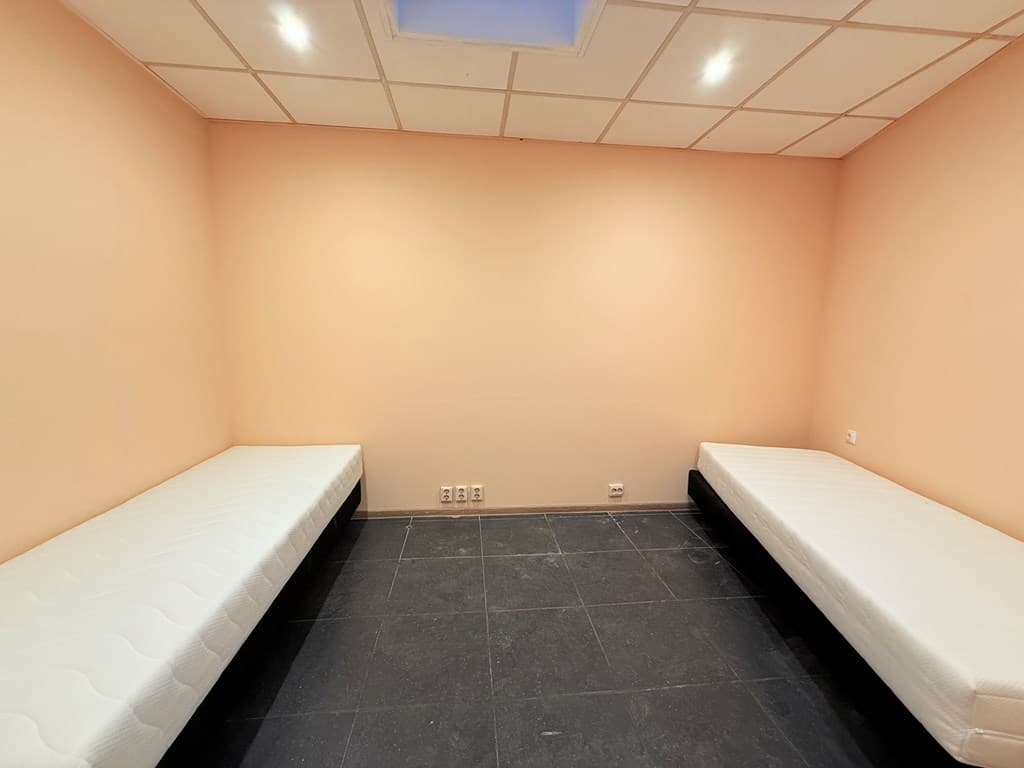 Twee eenpersoonsbedden naast elkaar in een lege kamer met een lichtgekleurde wand en een verlaagd plafond.
