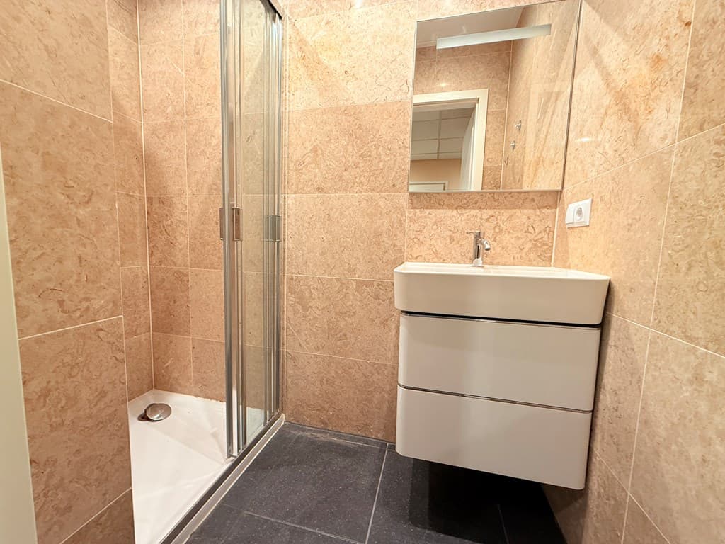 Douchecabine met glazen wanden en wastafel in een badkamer met marmerlook wandtegels en een donker tegeltapijt