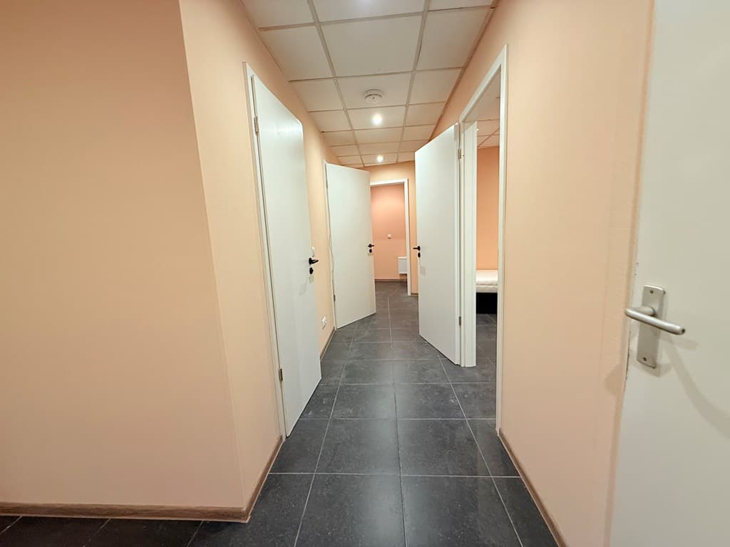 Een gang met lichtgekleurde muren en meerdere deuren, afgewerkt met donkere vloerbedekking.