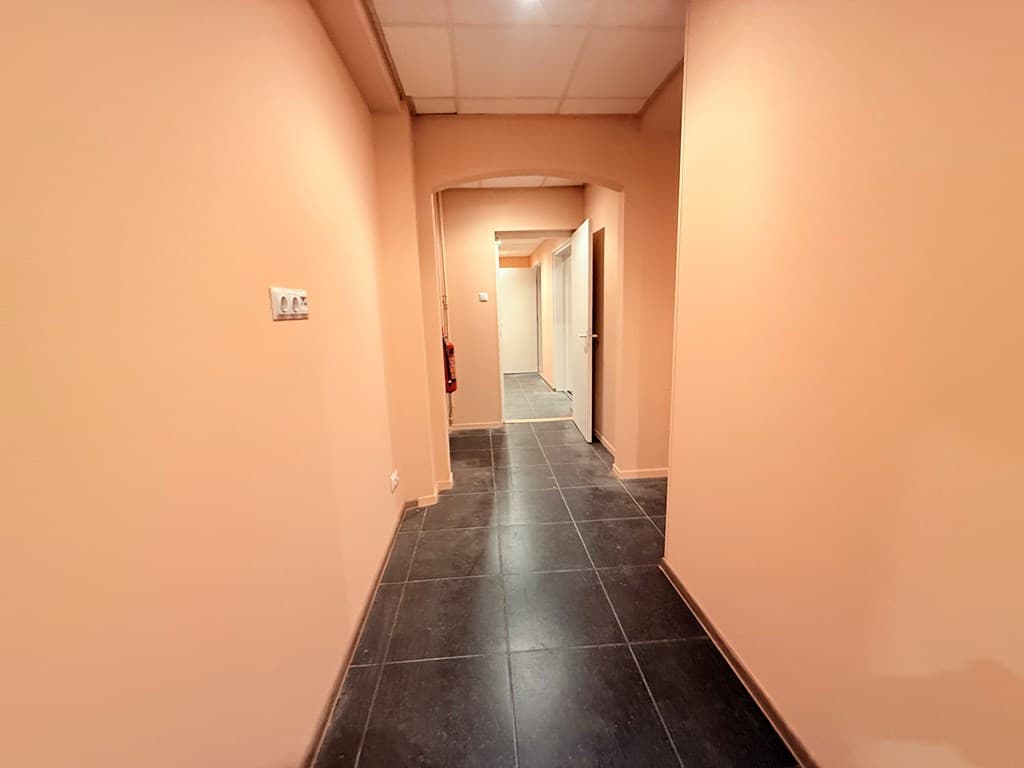 Een lange gang met lichtgekleurde muren en een donkere tegelvloer, met deuropeningen aan beide zijden.