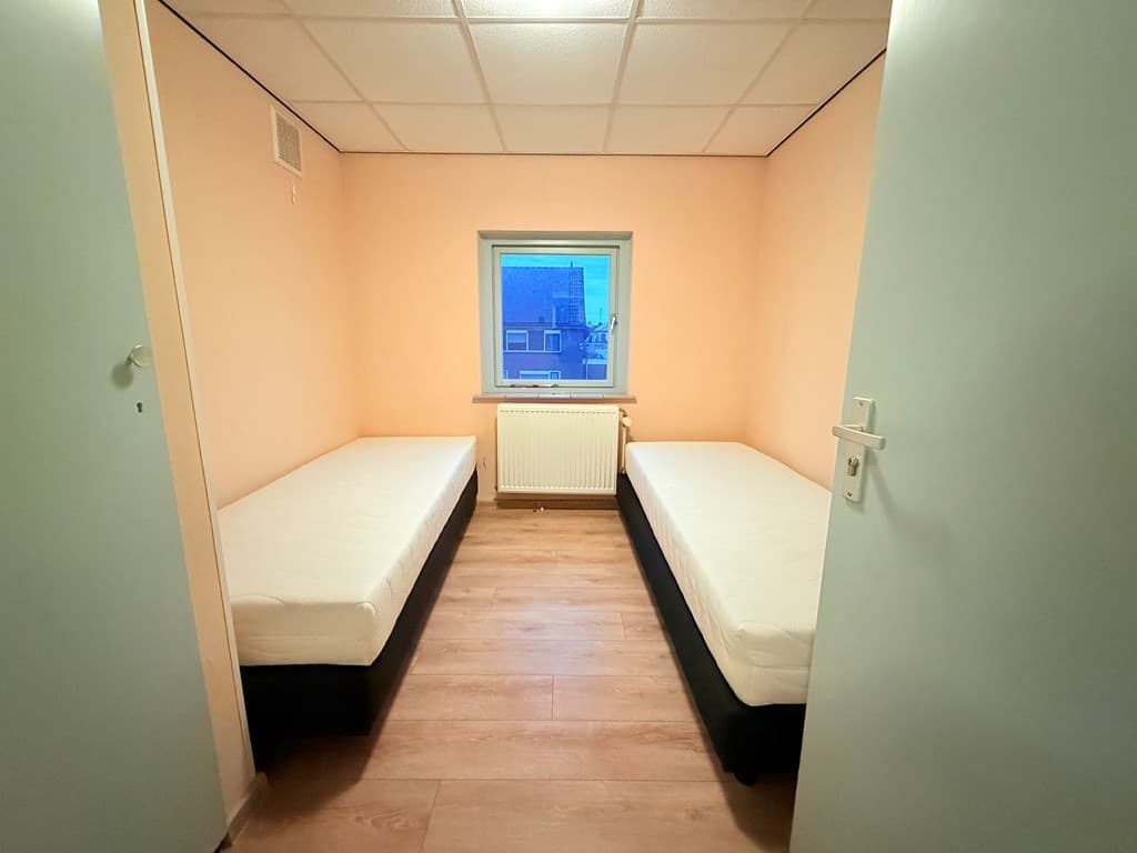 Slaapkamer met twee aparte bedden en een raam met uitzicht op de straat, muren in zachte pastelkleuren.