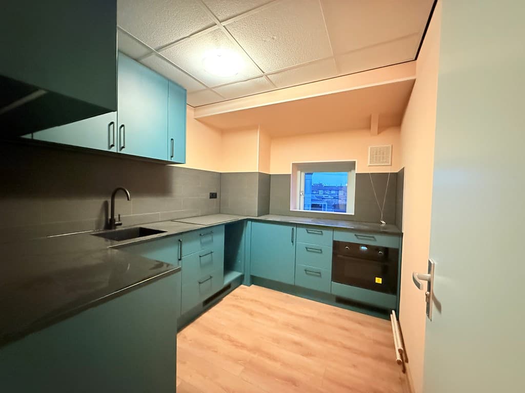 Keuken met blauwe kastjes, zwart aanrechtblad, donkere tegels en een raam met uitzicht op buitenlicht