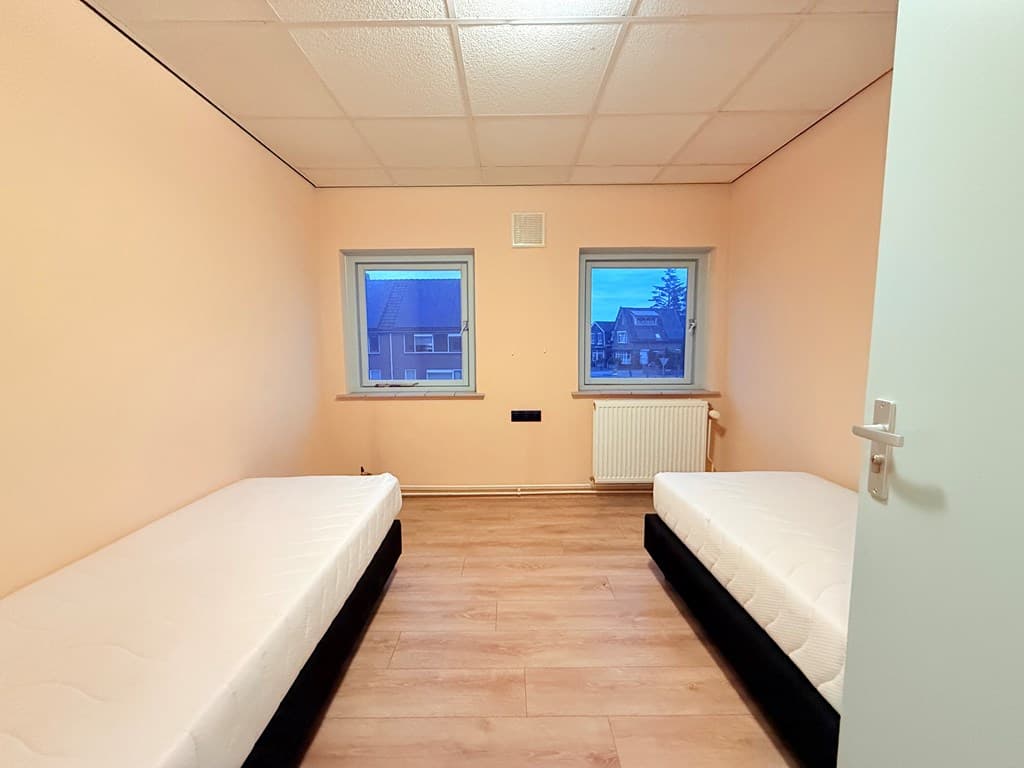 Slaapkamer met twee aparte bedden, lichtroze muren en ramen met uitzicht op de straat.