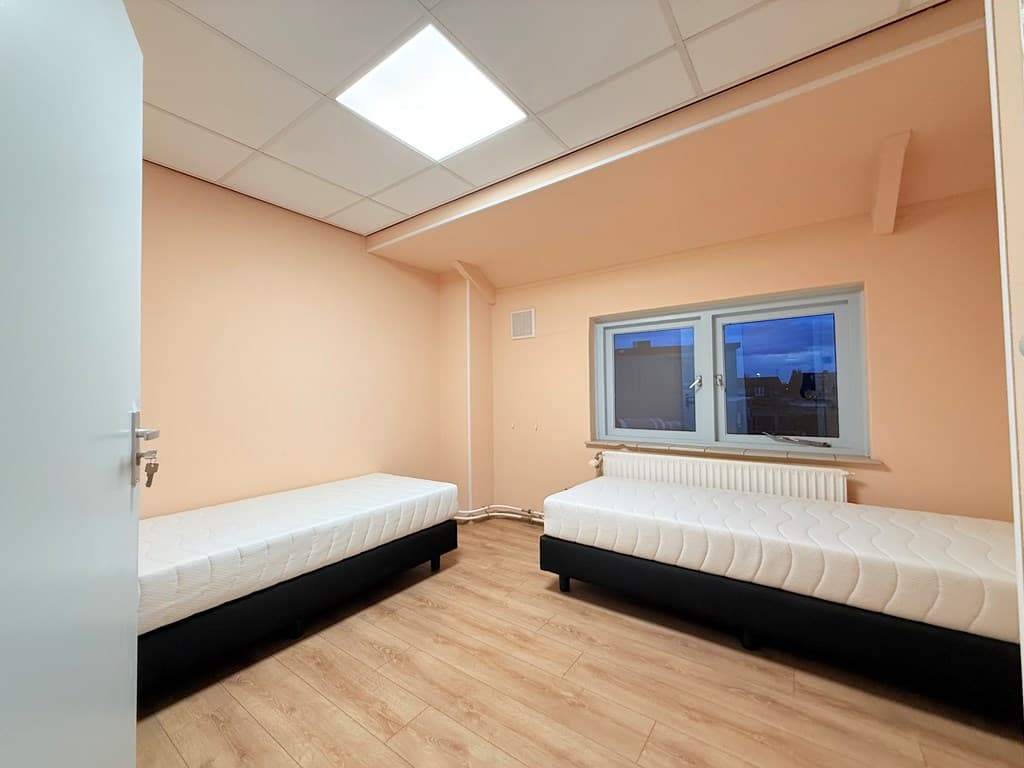 Slaapkamer met twee losse bedden, een raam en een beige muur op de achtergrond met houten vloer.