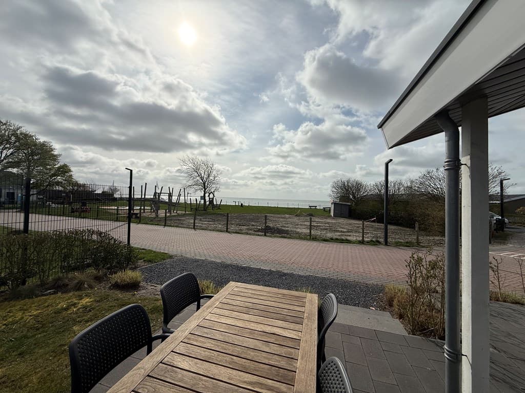 Uitzicht vanaf het terras met houten tafel en stoelen, grasveld en water op de achtergrond onder een bewolkte lucht