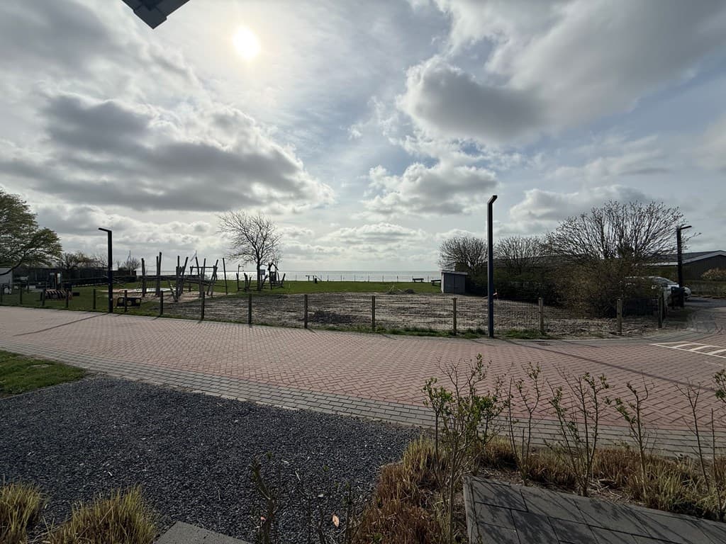Uitzicht op het grasveld met speeltoestellen en bomen bij EuroParcs Markermeer met een wolkenluch boven water
