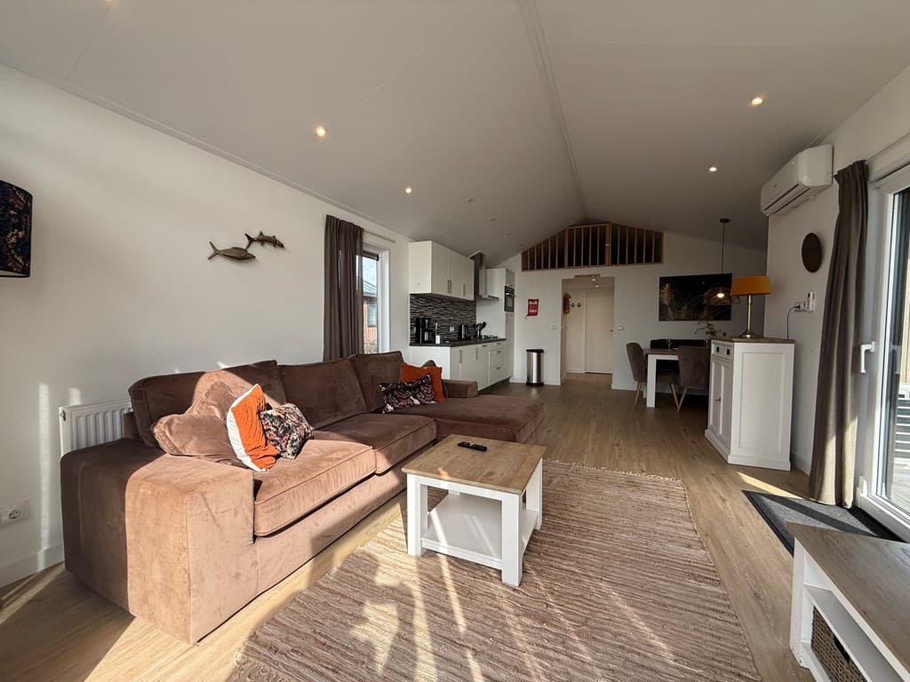 Lichte woonkamer met een bruine bank, witte kast, en open keuken met moderne inrichting en vloeren van hout.