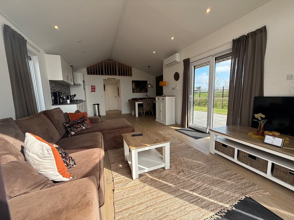 Woonkamer met een bruine bank, wit tv-meubel en openslaande deuren naar een terras met uitzicht op de natuur