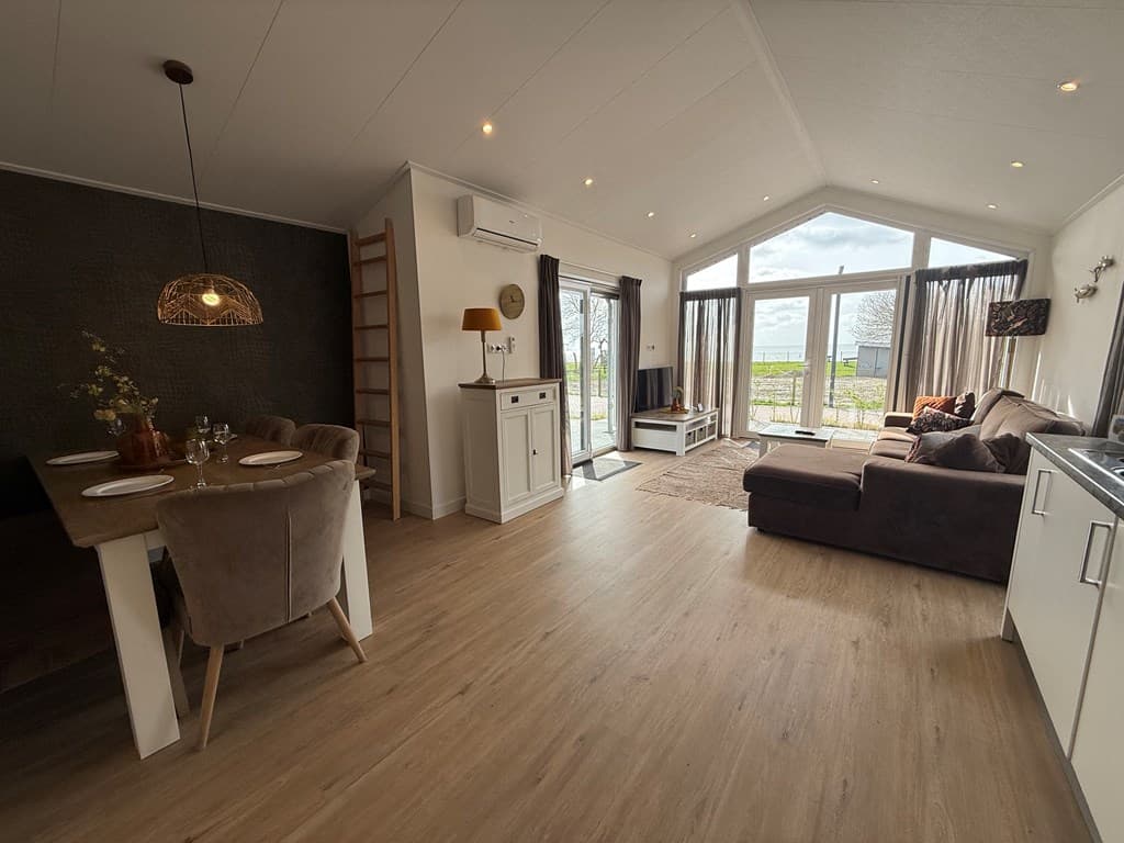 Licht interieur van een vakantiewoning met zithoek, eettafel en grote ramen met uitzicht op het buitengebied.