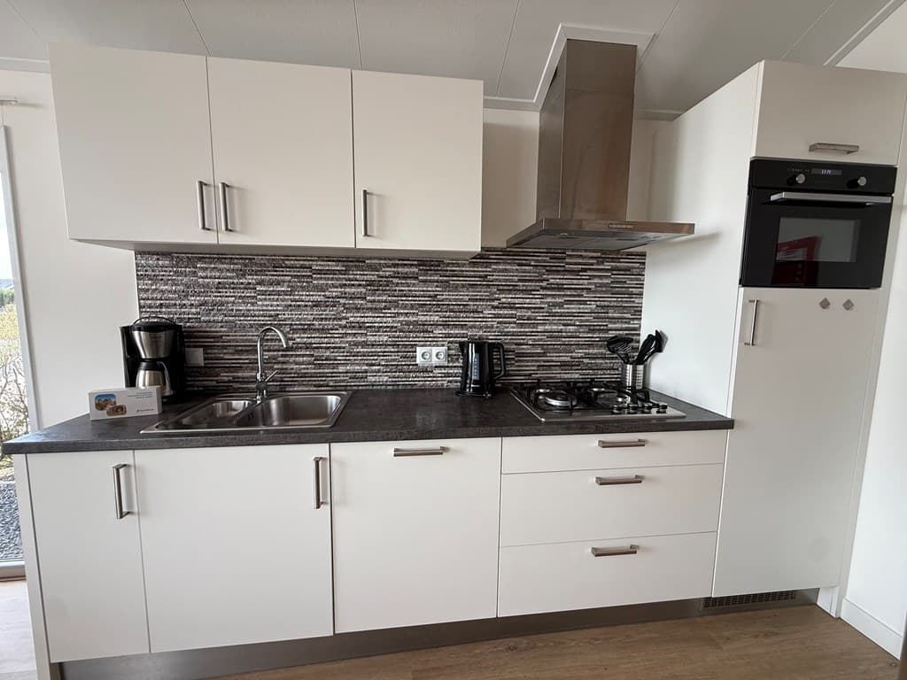Moderne keuken met witte kasten, een gasfornuis, oven, en een koffiezetapparaat, afgewerkt met een stenen achterwand.