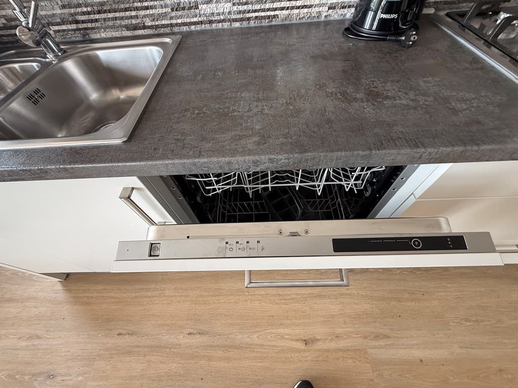 Keukenaanzicht met een open vaatwasser onder een aanrechtblad en een spoelbak, met een koffiezetapparaat op de achtergrond.