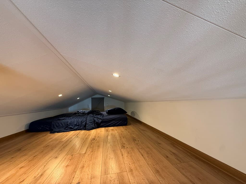 Zolderruimte met een laag plafond, houten vloer en een tweepersoonsbed met donkere beddengoed.