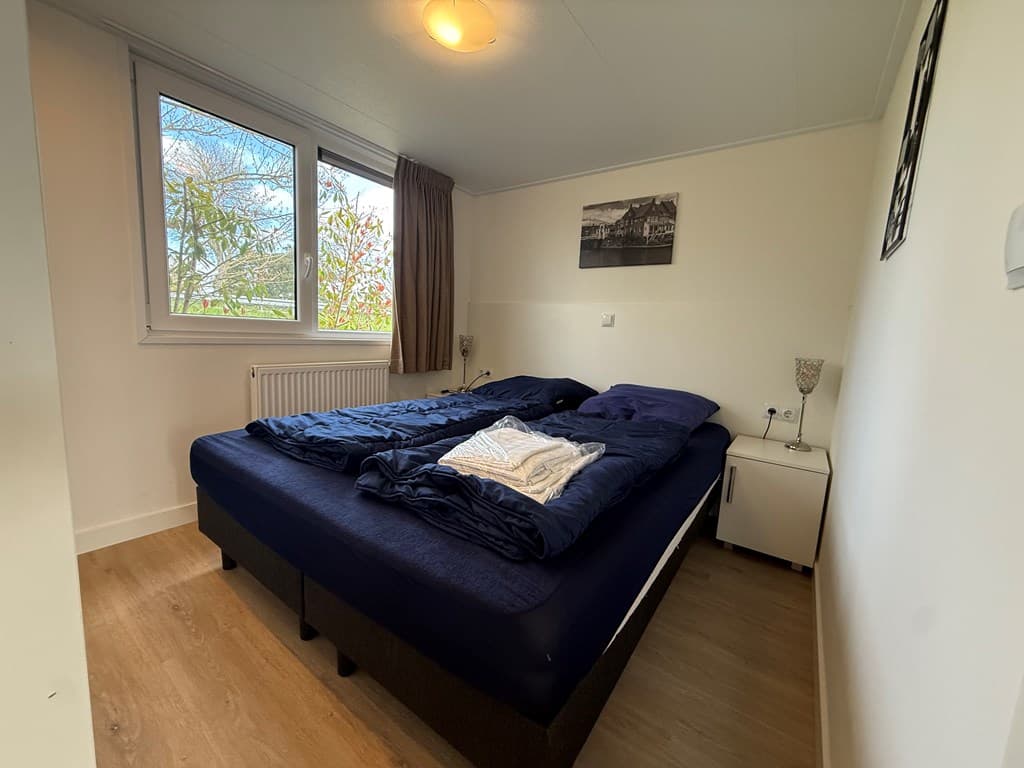 Slaapkamer met een tweepersoonsbed, nachtkastje en raam met uitzicht op groen.