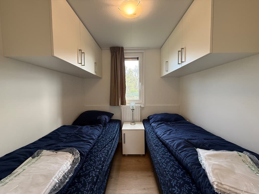 Slaapkamer met twee eenpersoonsbedden, nachtkastje en kastjes boven de bedden. Raam met gordijn zichtbaar.