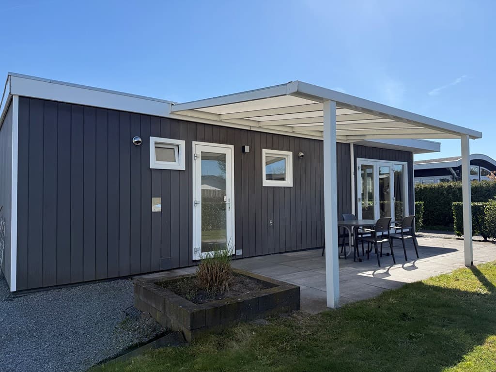 Moderne vakantiewoning met overdekt terras en tafel met stoelen, omringd door groen gras en struiken.