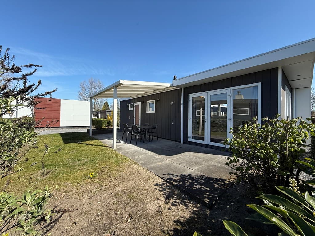 Moderne vakantiewoning met overdekt terras, tuin en ruime raampartijen onder een heldere blauwe lucht.