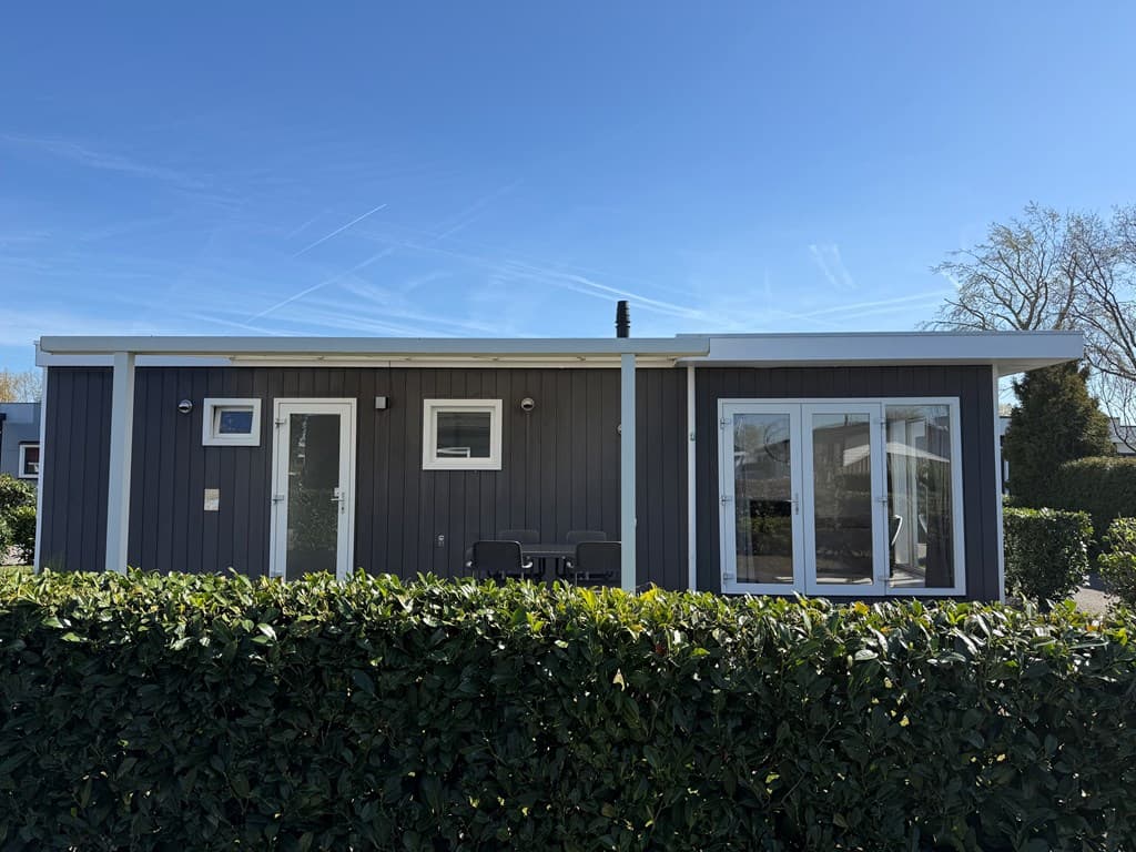 Moderne vakantiewoning met een houten gevel, grote ramen en een terras met stoelen, omgeven door groene heggen.