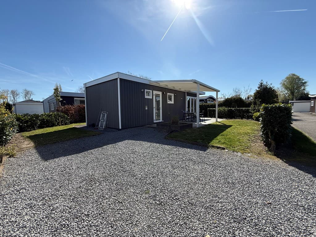 Moderne vakantiewoning met grijze gevel en overdekt terras, gelegen op een gravelperceel omringd door groen.