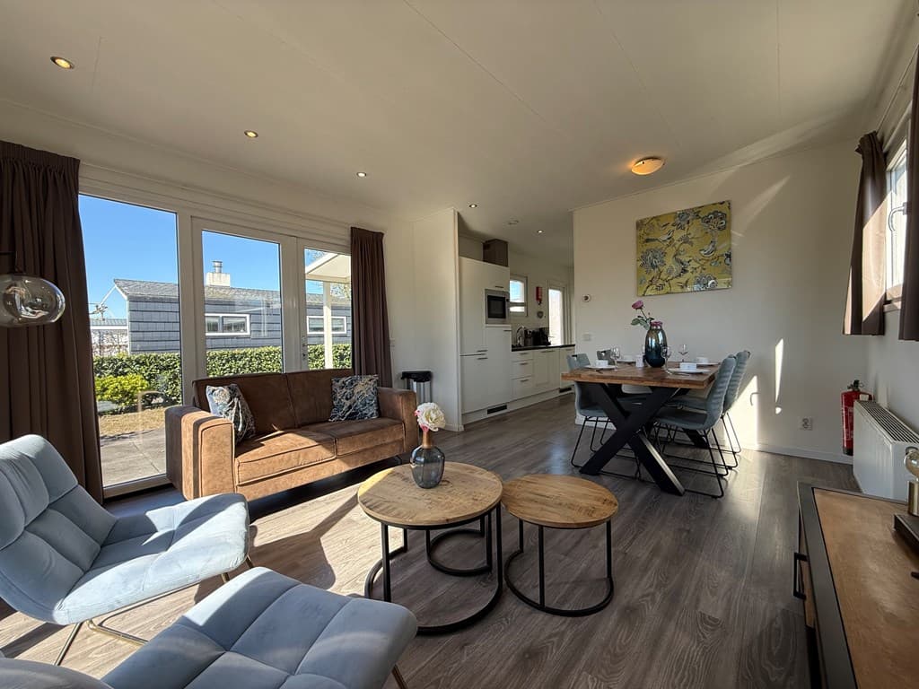 Lichte woonruimte met een bank, stoelen, eettafel en open keuken, grote ramen met uitzicht op de buitenruimte.