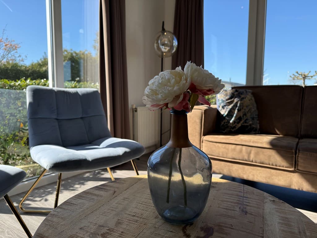 Lichte zithoek met een blauwe stoel, bruine bank en een vaas met bloemen op een houten tafel.