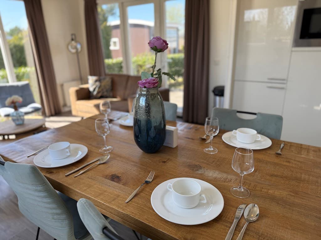 Eetkamertafel met servies en glazen, vaas met bloemen op de tafel, bank en grote ramen op de achtergrond.
