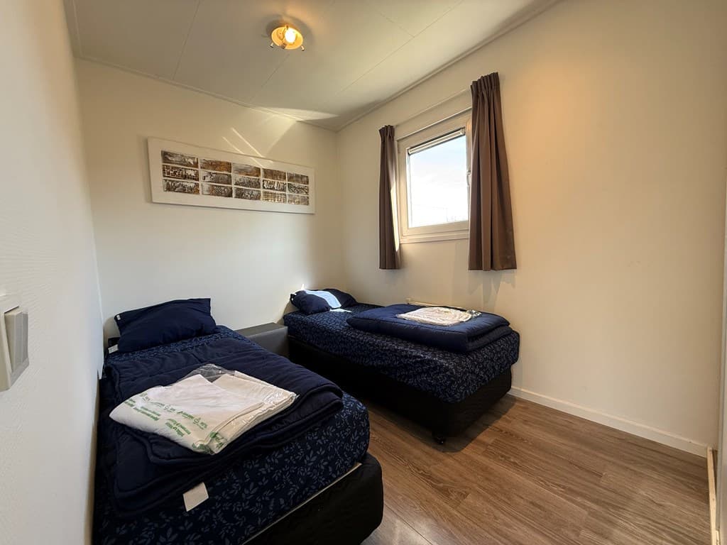 Slaapkamer met twee eenpersoonsbedden, gordijnen en raam met uitzicht op buiten, lichte muur en houten vloer.
