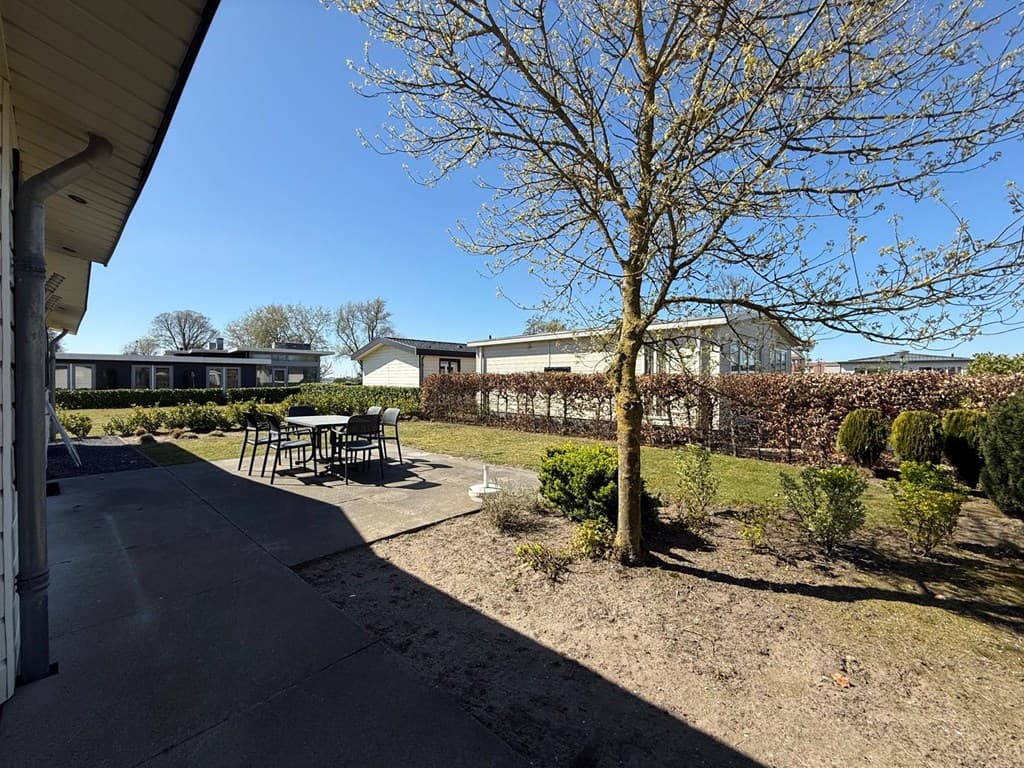 Tuin met gras en beplanting, terras met six-pack tafel en stoelen, zicht op naastliggende woningen en blauwe lucht.
