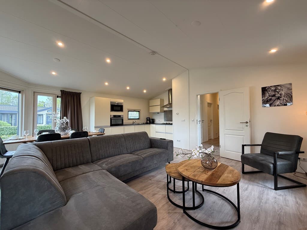 Ruime woonkamer met een moderne bank, salontafel, keuken en eethoek, grote ramen en lichte inrichting.