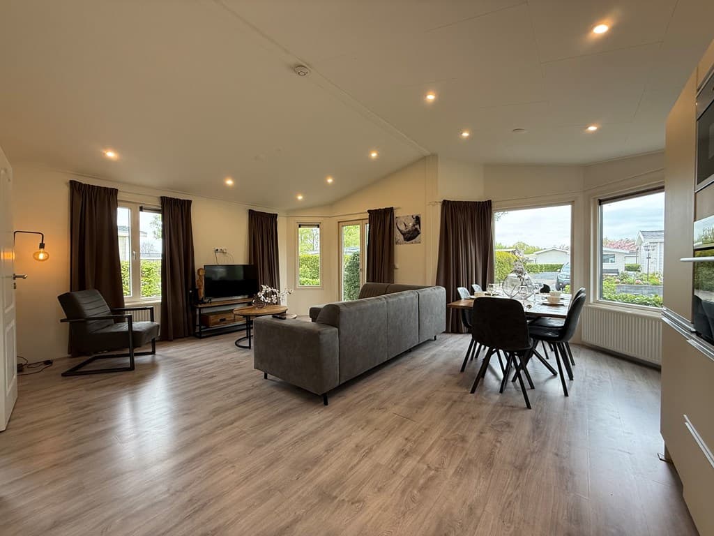 Ruime woonkamer met grote vensters, een zithoek met bank en stoel, en een eettafel met stoelen bij de ramen.