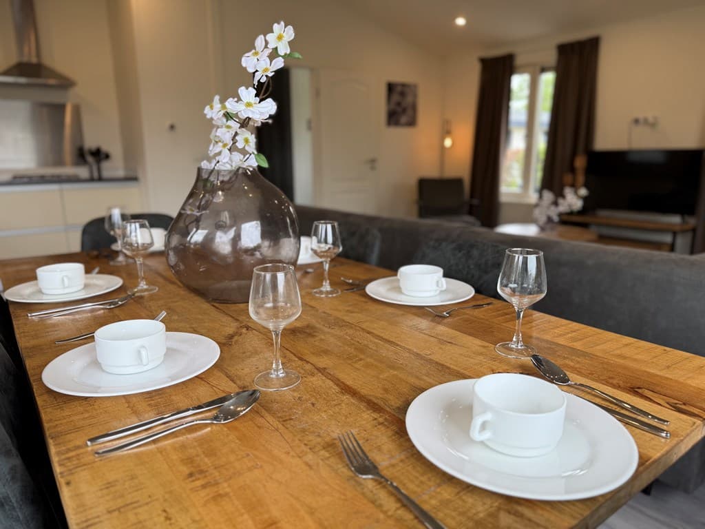 Eettafel met zes borden, glazen en bestek, een grote vaas met bloemen en een open woonkamer met moderne inrichting.