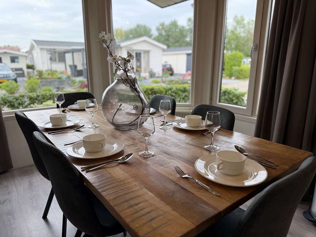 Eettafel met servies en glazen, uitzicht op de buitenomgeving met vakantiewoningen en groenvoorziening.