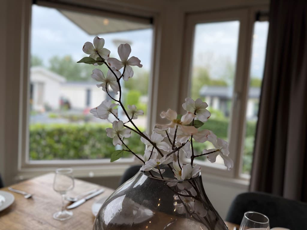 Tafel met een vaas met takken en bloemen, uitzicht op buiten met bomen en vakantiewoningen door de ramen.