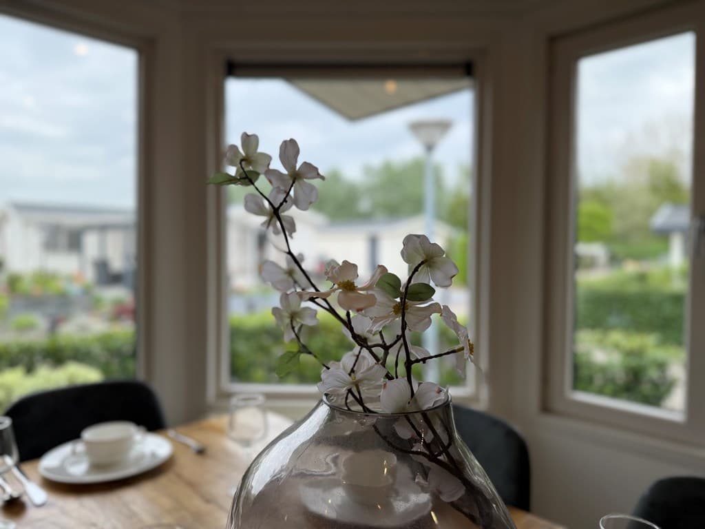 Vaasje met takken met bloemen op een houten tafel, uitzicht op groene omgeving door grote ramen met zicht op vakantiehuisjes