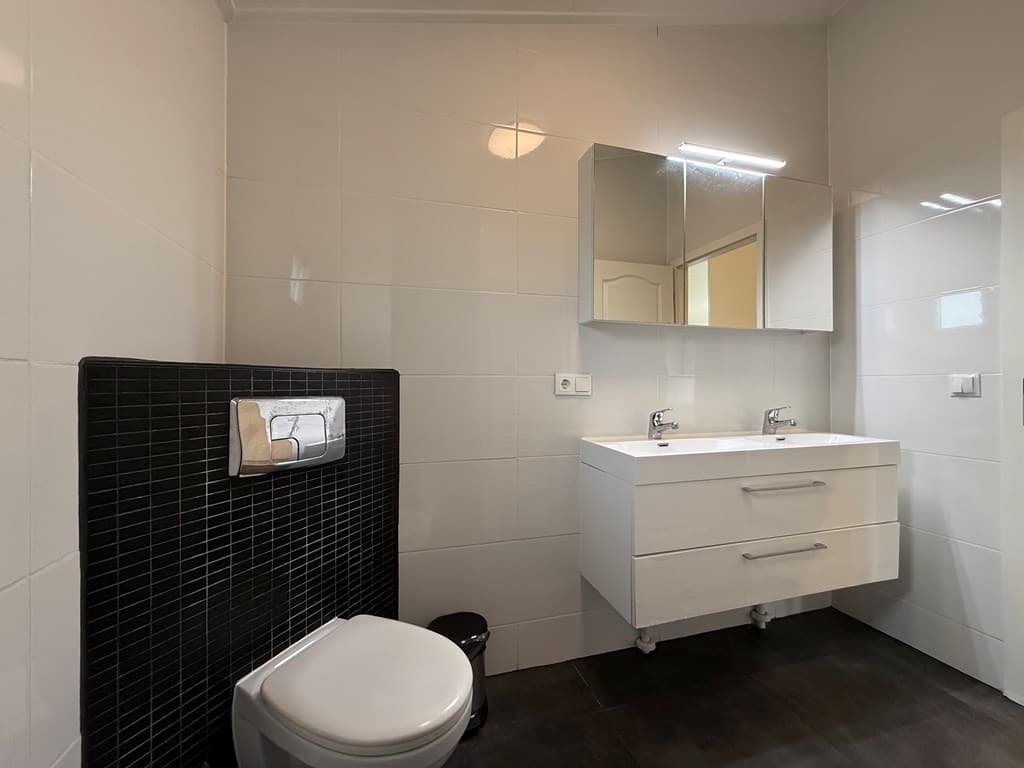Moderne badkamer met witte wandtegels, zwart tegelwerk en dubbele wastafel met spiegels boven de wastafel.