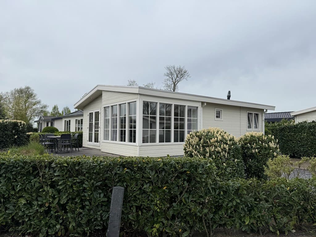 Moderne vakantiewoning met grote glazen ramen en terras, omringd door groene hagen en struiken.