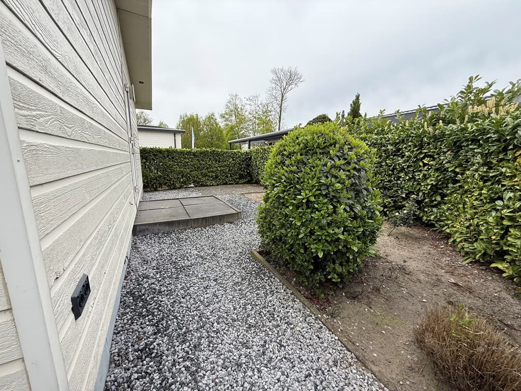 Zijaanzicht van de woning met grind en een verharding, omgeven door groene hagen en struiken.