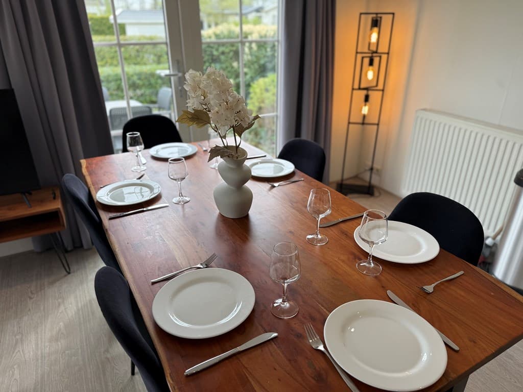 Eethoek met houten tafel, zes borden, glazen en een vaas met bloemen bij de ramen met uitzicht op de tuin